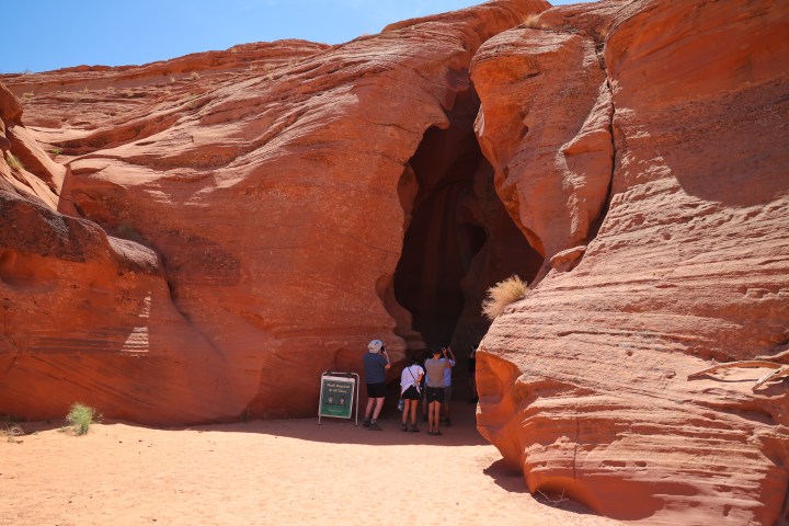 People at a red sandstone cave entrance under a clear blue sky.