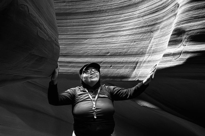 Person stands between curved rock formations in a canyon, looking up, with light illuminating the scene.